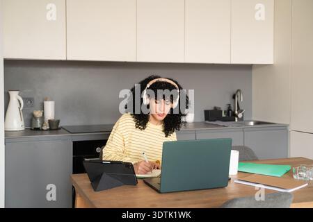 A woman wearing headphones works remotely in a modern kitchen. She is writing notes in a notebook while using a laptop, with a tablet and other items Stock Photo