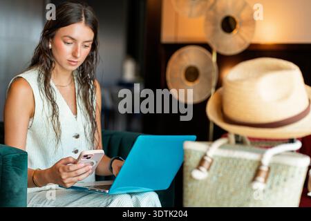 A woman in a hotel lobby multitasks using a smartphone and laptop. The serene setting with a stylish hat and bag nearby suggests a blend of work and l Stock Photo