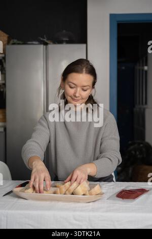 A woman in a gray sweater arranges slices of bread on a tray in a modern kitchen. She appears focused and content as she carefully prepares the items Stock Photo