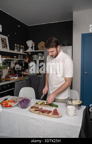 A couple is seen actively preparing a meal in a cozy kitchen. The man slices cucumbers while the woman focuses on tasks by the counter, surrounded by Stock Photo