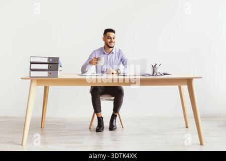 Man Sits at Desk Smiling While Holding Coffee Cup and Working on Laptop in an Office Setting Stock Photo