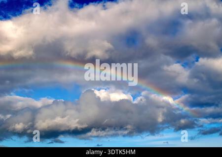 A vibrant rainbow arcs across a dynamic sky filled with a mix of fluffy white and dark grey clouds, with patches of bright blue sky visible. The natur Stock Photo