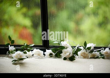 A close-up shot of a white rose garland with green leaves draped along a window sill. The window pane is covered in raindrops, creating streaks and a Stock Photo