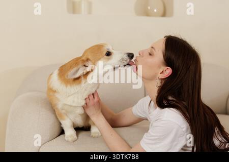 Young woman sitting on a sofa gently holding her corgi while the dog licks her face. Warm domestic moment showing affection, trust and joyful companio Stock Photo
