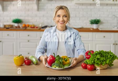 Portrait of happy smiling young woman sitting at kitchen table with fresh vegetables Stock Photo