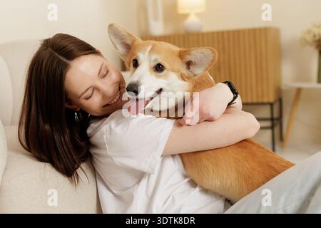 Smiling woman embracing her corgi dog while relaxing on sofa in a cozy living room. Warm indoor lifestyle scene showing affection, companionship and e Stock Photo