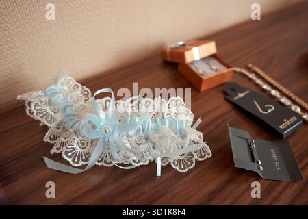 A beautifully crafted white lace garter, adorned with light blue satin ribbons and a sparkling embellishment, lies on a warm wooden surface. This trad Stock Photo