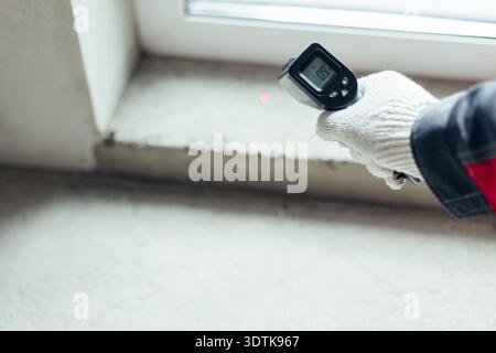 Hand In Glove Holding Infrared Thermometer With Laser Dot On Window Sill. Temperature Measurement In Construction Setting. Stock Photo