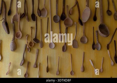A vibrant wall display of numerous handmade wooden spoons in various shapes, sizes, and shades of brown, hung vertically on a bright yellow background Stock Photo