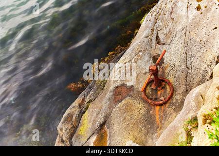 A high-angle view captures a weathered, rusty metal mooring ring firmly embedded into a light-colored, textured rock face along a coastline. Streaks o Stock Photo