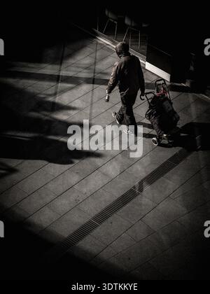 High angle view of an unrecognizable person pulling a trolley through a dramatic shaft of sunlight and deep shadows on a tiled urban pavement. Stock Photo