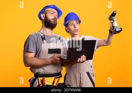 Caucasian coworkers discuss renovation plans, comparing measurements with digital data on laptop. Female supervisor gestures toward spot with drill in hand as male employee listens and holds device. Stock Photo