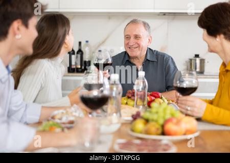 Senior man at kitchen table with wife, grown son and daughter, enjoying conversation at festive table with wine and snacks Stock Photo