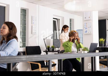 Overworked female employee rubbing eyes in pain, showing stress and mental fatigue from demanding corporate workday. African American employee sits stressed at laptop, trying to regain focus. Stock Photo