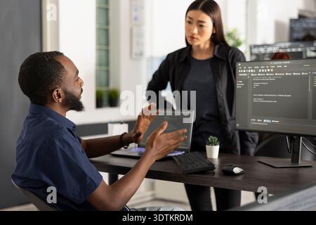 Manager reviews application progress as coworker presents updated software code information. The discussion takes place beside computer, highlighting collaboration and planning in startup office. Stock Photo