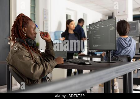African american developer thinking while working on app development at her desk, surrounded by diverse colleagues in open tech workplace. Female programmer pondering on solutions to improve website. Stock Photo