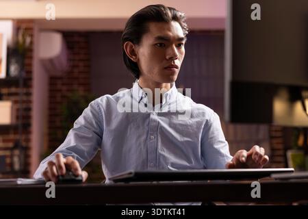 Young male entrepreneur typing and reviewing strategy plans on desktop pc, dedicated on achieving business goals. Asian man works from home at night, completing unfinished tasks and reaching deadlines Stock Photo