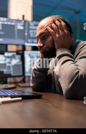Overworked programmer rests head in palms during difficult coding session. Male developer analyzes complex data on laptop with exhausted expression, represents programming stress and technical pressure Stock Photo