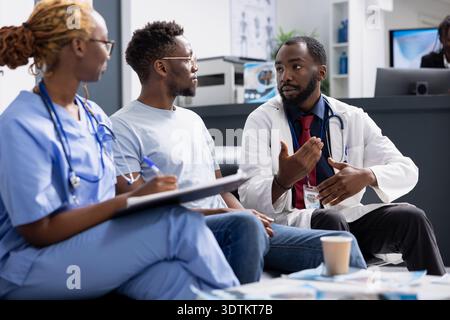 Black male doctor in lab coat speaks with patient during clinic appointment, reviewing documents and discussing health concerns. Female nurse takes notes as physician explains treatment information. Stock Photo