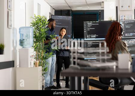 Diverse team of professional coders discuss programming solutions and review source code on laptop. African American and Asian developers support teamwork and digital technology growth. Stock Photo