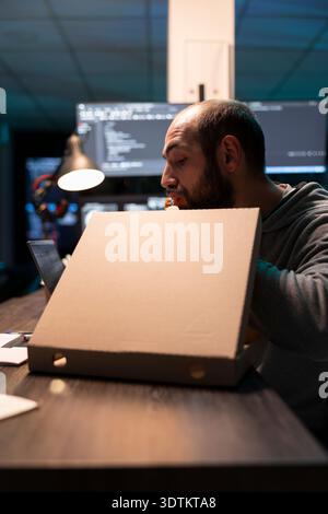 Software developer eating pizza straight from box and analyzing decoding information on laptop. Screens filled with code surrounds programmer, representing busy work routine and late hour productivity Stock Photo