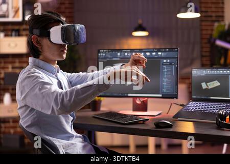 Young man wearing vr headset and interacting with 3d cad software on desktop and laptop screens. Asian male individual developing container model with ar technology in home office during evening hours Stock Photo