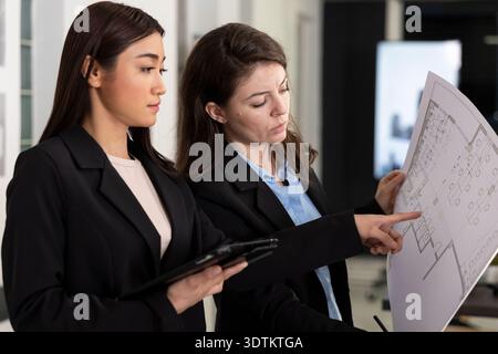 Diverse coworkers analyze architectural drawings, looking at floor plan details and structural elements. Asian and caucasian architects reviewing building measurements and taking notes on tablet. Stock Photo