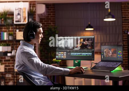 Young man focuses on matching audio with video footage during post production. Asian male filmmaker enjoys music during editing process on digital devices, working from cozy home office studio. Stock Photo