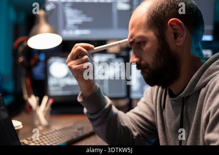 Exhausted tech freelancer leans forward holding pen to forehead while reviewing coding errors. Sad male programmer feeling fatigued and stressed after hours of intense programming work. Stock Photo