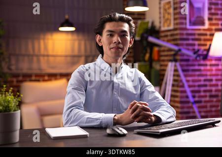 Confident professional man and entrepreneur sits at desk with computer keyboard, looking at camera. Portrait of focused asian male executive and leader working remotely in modern apartment. Stock Photo