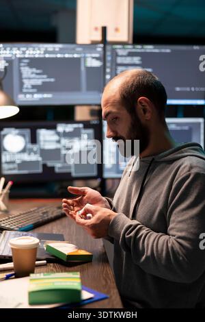 Male IT worker holds medication at desk, managing fatigue after long hours solving technical problems. Caucasian programmer checks pills while surrounded by monitors displaying complex system code. Stock Photo