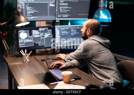 Male developer leans toward several monitors while reviewing complex lines of code during late night work session. Concentrated software engineer works in tech office, refining tasks with precision. Stock Photo