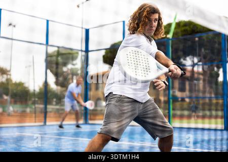 Focused middle-aged Latin man playing padel on court. View through tennis net Stock Photo