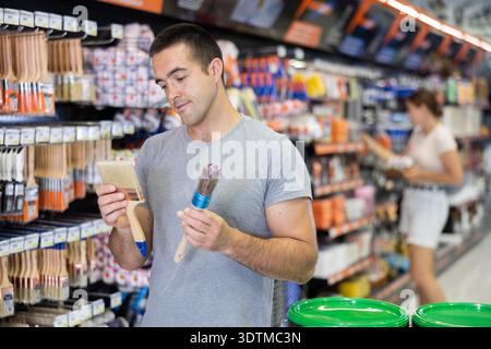 Focused young man choosing brushes in painting supplies store Stock Photo