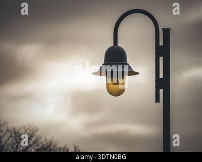 Single streetlamp with yellow glass bulb illuminated against a cloudy sky on a moody overcast day Stock Photo
