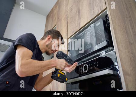 Man in workwear installing built in microwave above oven using cordless screwdriver in wooden kitchen cabinet Stock Photo