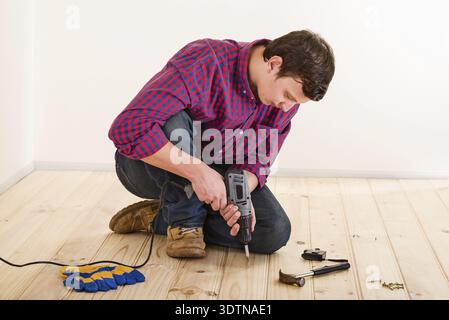 Young worker with screwdriver installing wooden flooring in the room Stock Photo