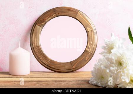 Round blank wooden frame with natural finish rests on wooden table beside pink candle and white flowers set against soft pink wall, creating beautiful Stock Photo
