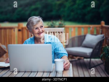 Mature woman sitting on balcony with laptop, working remotely. Stock Photo