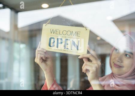 A young female shop owner hangs an open sign, symbolizing small business operation, entrepreneurship, and welcoming customers with confidence. Stock Photo