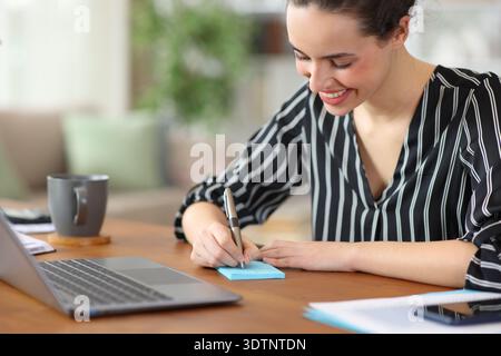 Happy freelance worker writing with pen in blue stiky note working at home Stock Photo