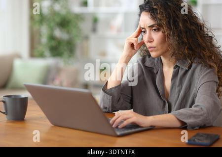 Worried adult woman at home checking bad news on laptop on a wooden table Stock Photo
