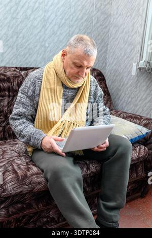 Senior man with gray hair sits on couch, wearing yellow scarf and sweater, looking concerned while using a laptop Stock Photo