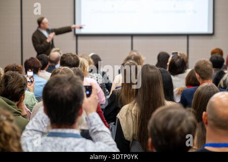 Crowd of professionals listening to presenter during business conference, some attendees recording slides with smartphones. Stock Photo