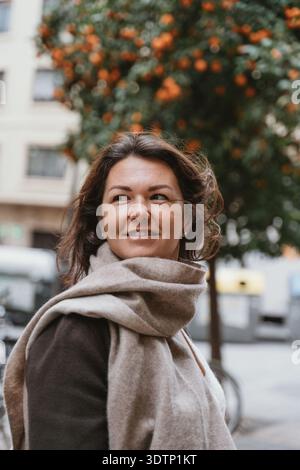 Woman in cozy scarf smiling outdoors with orange trees and buildings in the background. Hair gently blown by the wind, creating a serene and confident Stock Photo