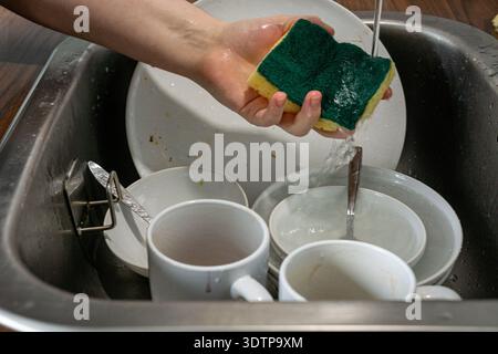 Close up of a hand squeezing a kitchen sponge while washing dirty dishes in a stainless steel sink. Stock Photo