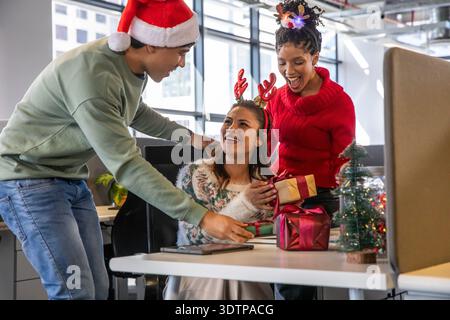 Diverse coworkers exchanging wrapped holiday gifts at office desk with Santa hat and tree Stock Photo