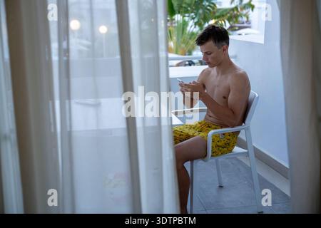 Young Man Using Smartphone While Sitting on Balcony in Summer Vacation Setting Stock Photo