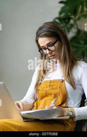 Focused woman consultant in overalls wear headphones using laptop, communicating with clients, makes a report on paper documents, works from home garden. Cozy office workplace, remote work || Model released Stock Photo