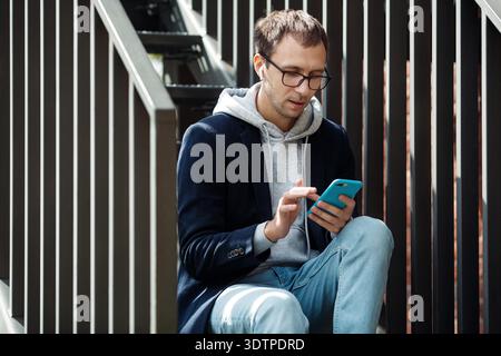 Casual guy in earphones browsing in smartphone sit on stairs outdoors. Freelancer entrepreneur read email. Office worker check social media on break outside listening to music with wireless headphones || Model released Stock Photo